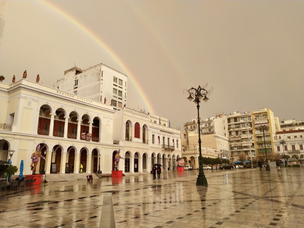 The Apollo Theatre at a wet King George Square in Patra, with big carnival puppet puppets and a double rainbow in the sky, photo by Sophie Huijts