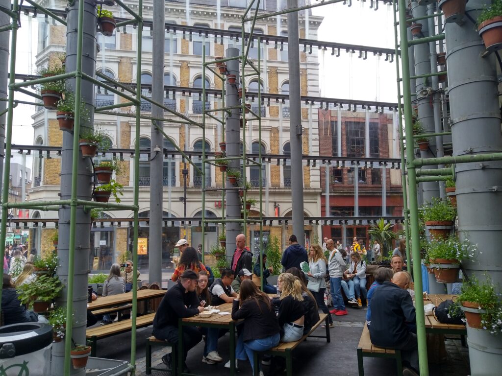 Picknick area with tables and benches inside Borough Market, with plants along pillars and old houses in the back, photo by Sophie Huijts