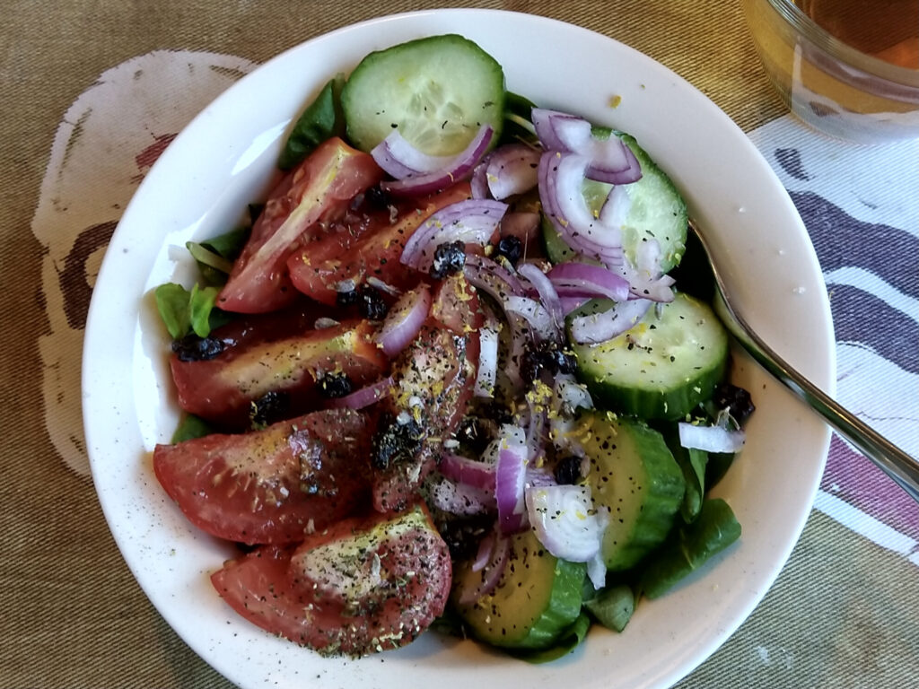 Bowl of salad with lettuce leaves, tomatoes, cucumber, red onions, currants and topped with herbs and lemon zest, prepared and photo by Sophie Huijts