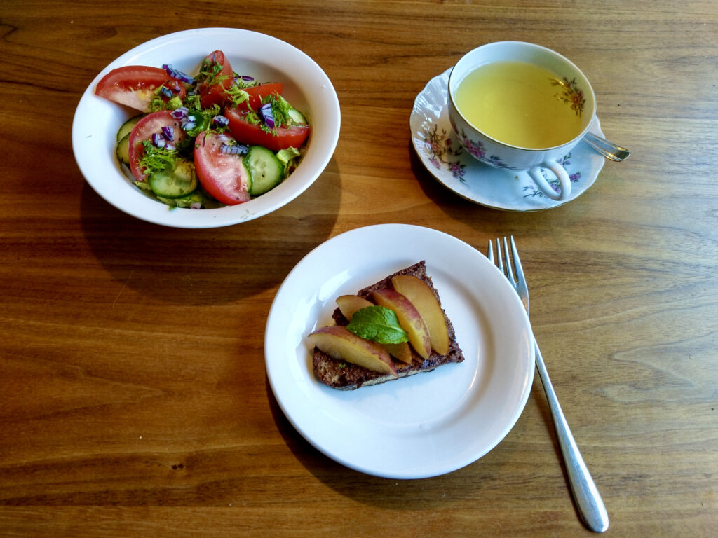 Breakfast table for one with a tomato cucumber salad, a cup of tea and a small plate with a piece of pain perdu topped with fresh plum and a mint leave, prepared and photo by Sophie Huijts